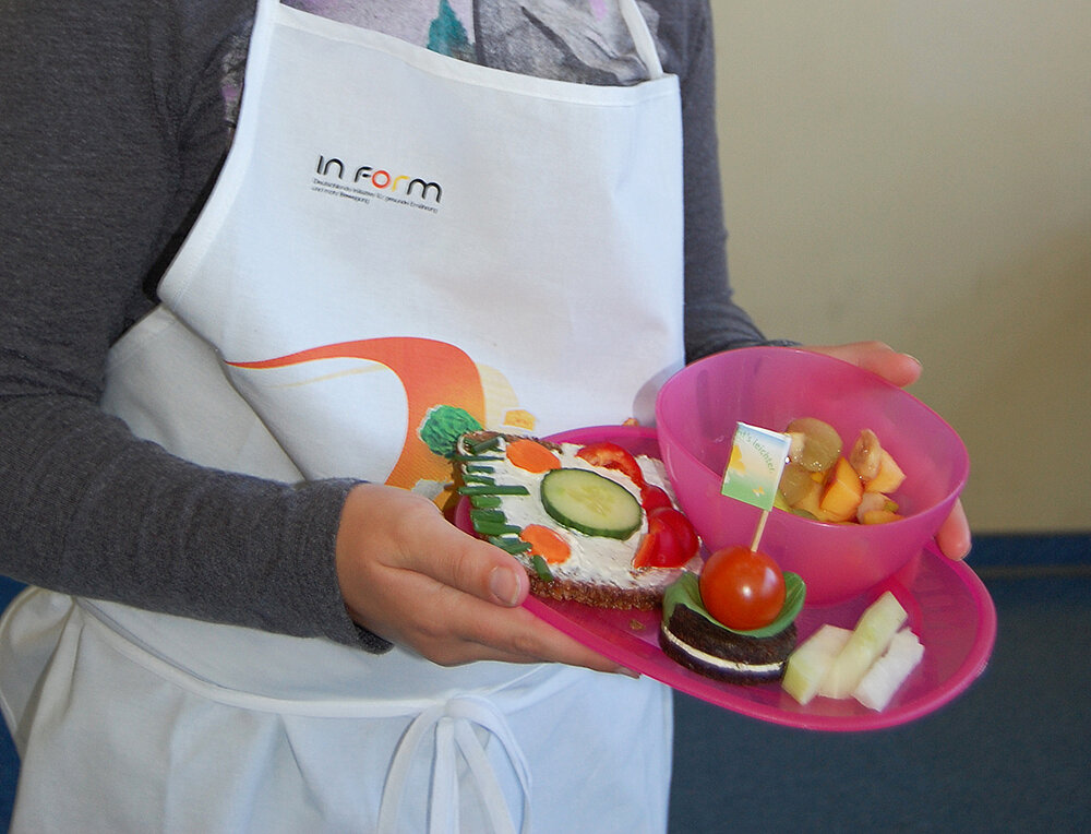 Ein Mädchen hält einen Teller mit einem Brotgesicht und verschiedenem Obst und Gemüse in der Hand.