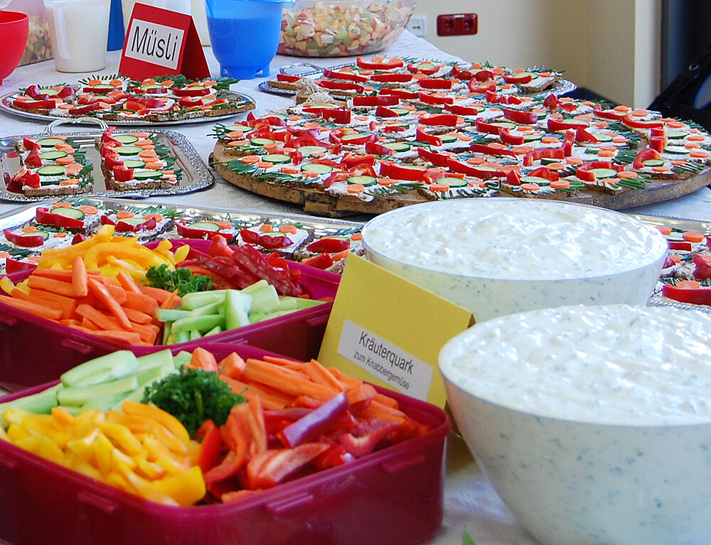 Auf einen Tisch stehen mehreren Schüsseln gefüllt mit Obst, Gemüse und Quark, Teller mit Brotgesichtern und weitere gesunde Leckereien