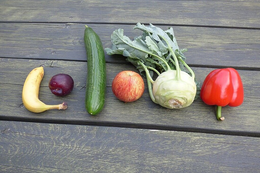 zum Europäischen Schulprogramm verschiedene Obst- und Gemüsesorten liegen in einer Reihe auf einen grünen Holztisch (1 Banane, 1 Pflaume, 1 Gurke, 1 Apfel, 1 Kohlrabi, 1 rote Paprika)