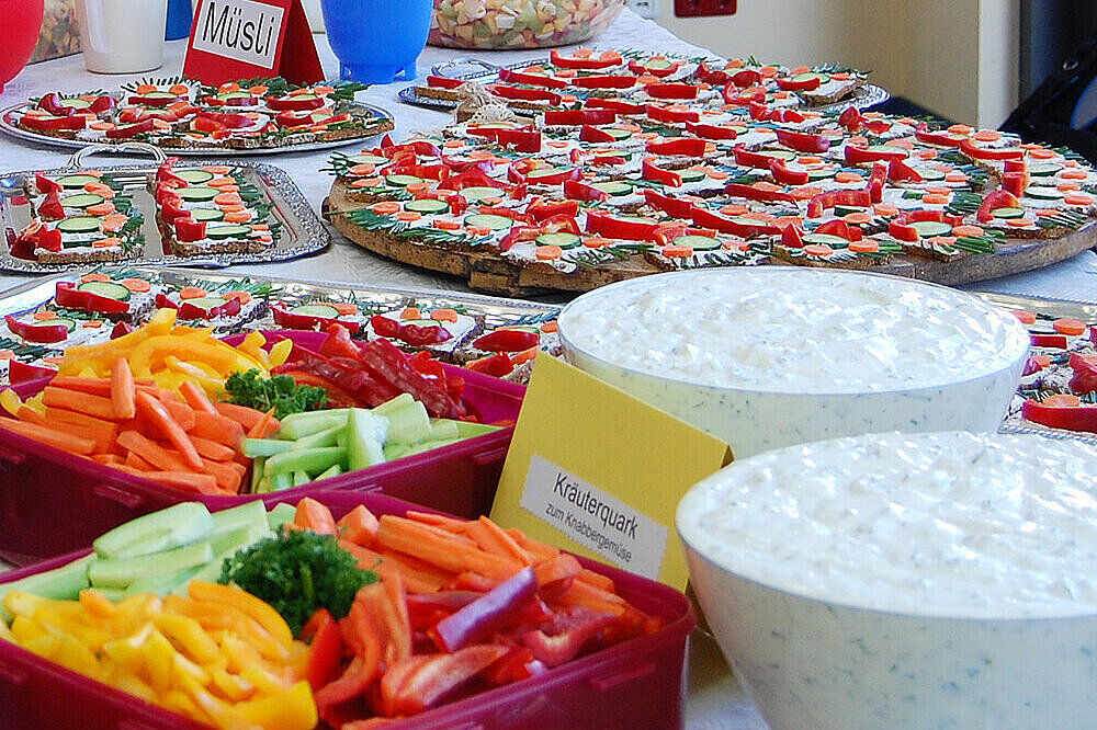 Zur Kita- und Schulverpflegung Auf einen Tisch stehen mehreren Schüsseln gefüllt mit Obst, Gemüse und Quark, Teller mit Brotgesichtern und weitere gesunde Leckereien